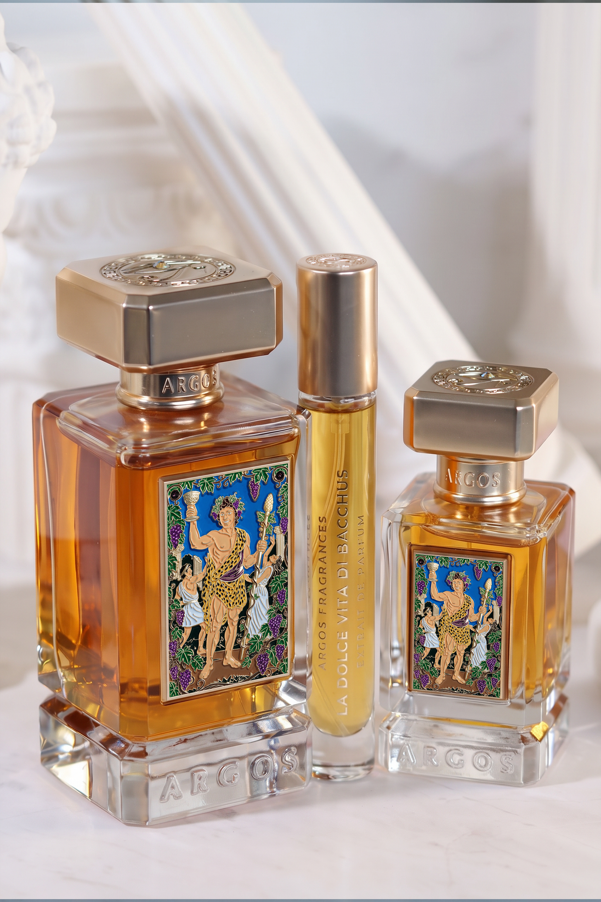 Three argos la dolce vita di bacchus perfume bottles displayed together on a light marble surface with a white draped background. The set includes a large square bottle, a tall cylindrical travel spray, and a smaller square bottle, all filled with amber liquid and featuring gold caps and a colorful classical illustration label.