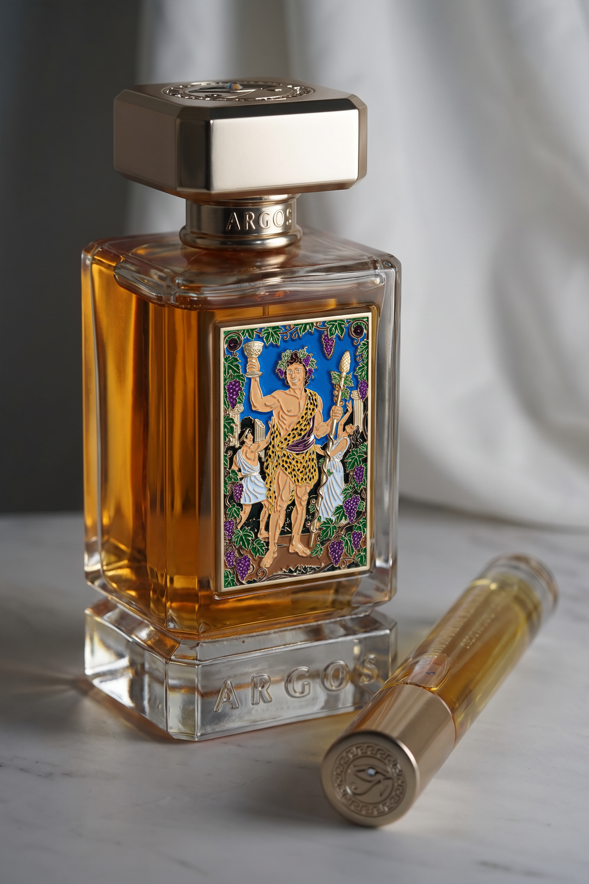 Argos perfume bottle filled with amber liquid, featuring a colorful Art Nouveau-style label and a gold-toned cap, shown on a marble surface with a matching cylindrical vial beside it.