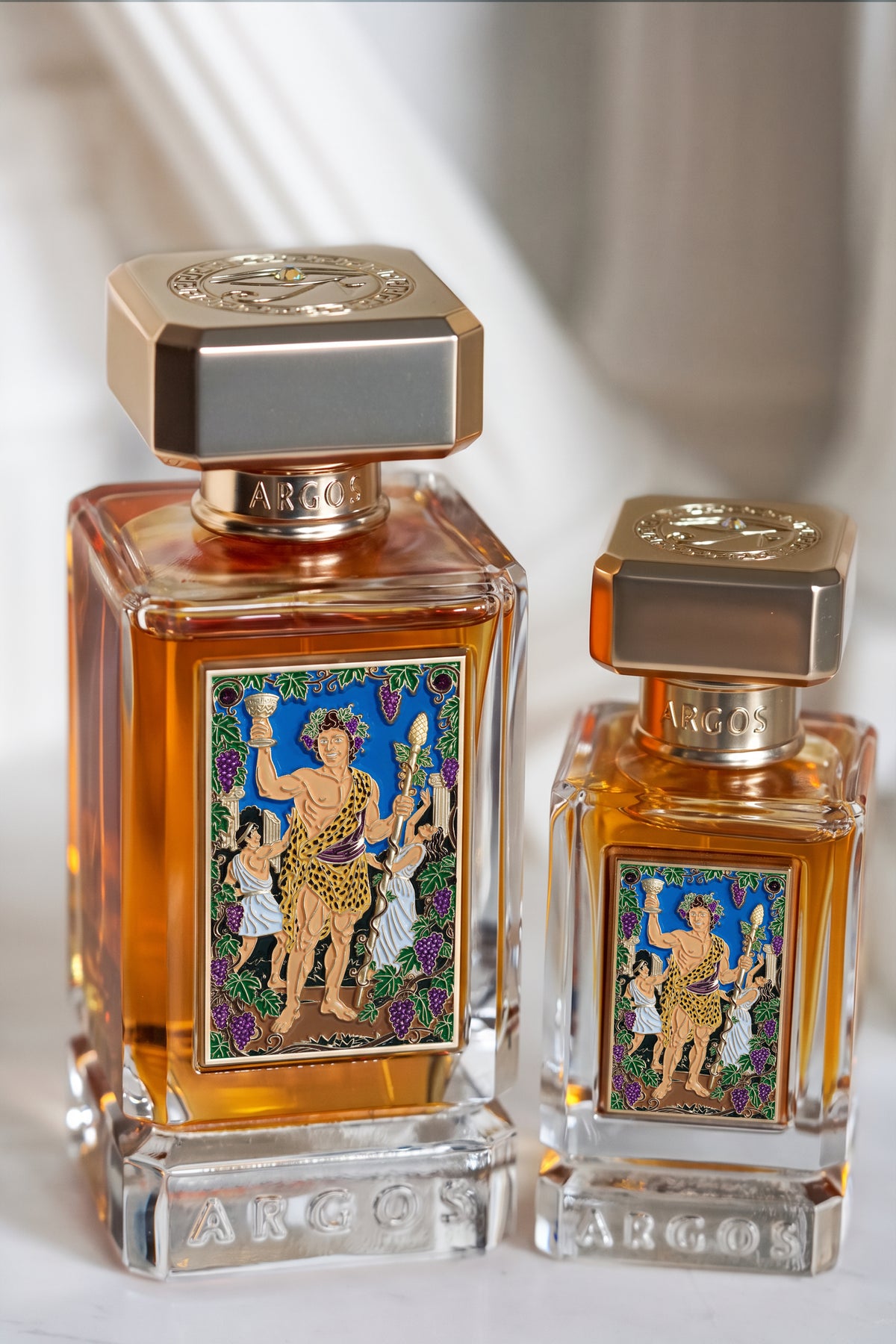 Two Argos perfume bottles with ornate gold caps and colorful illustrated labels, shown on a light marble surface with a soft blurred background.