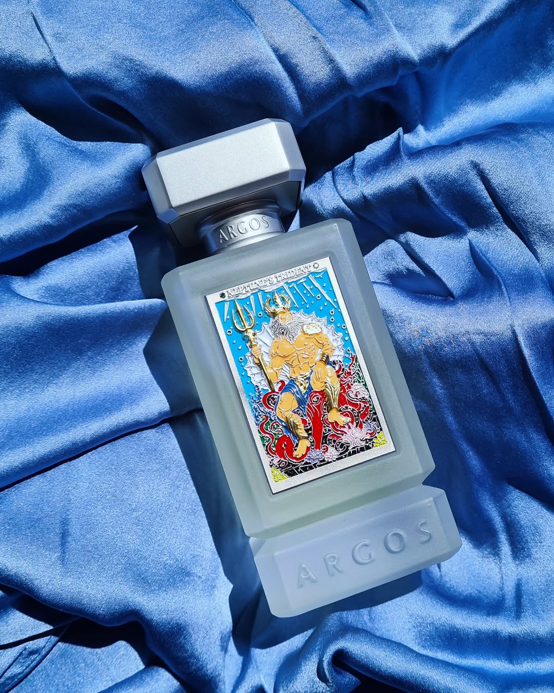 Argos Neptune's Trident Extrait de Parfum 100ml bottle with an illustrated Neptune label, placed on layered blue silk background.