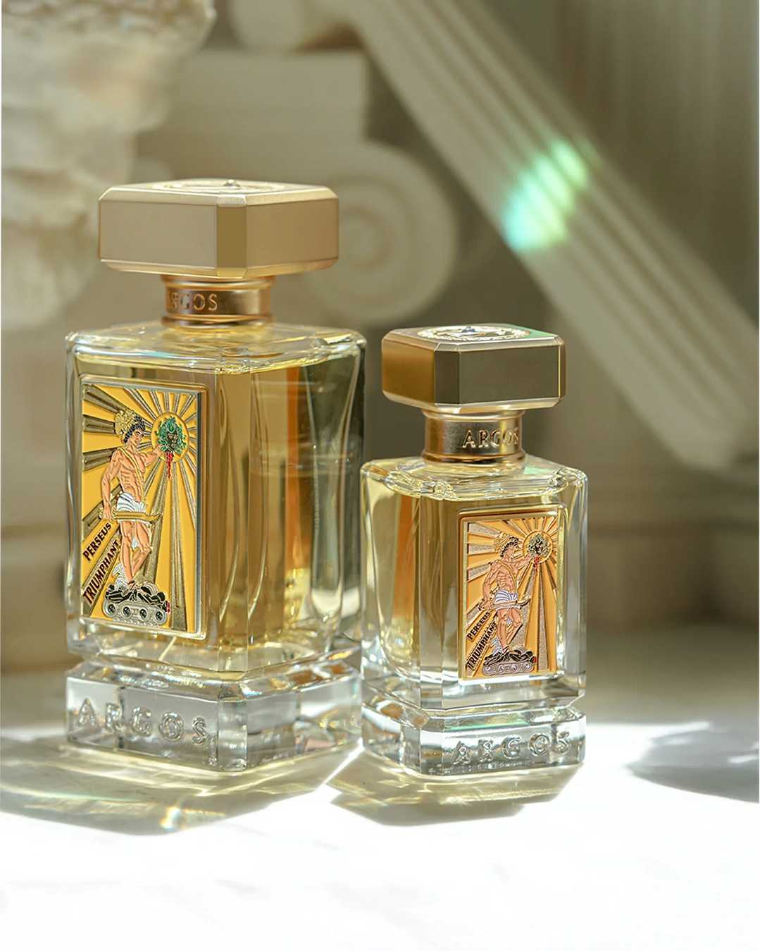 Two Argos Perseus Triumphant Extrait de Parfum bottles (100ml and 30ml) with gold accents and illustrated labels on a reflective surface, set against a Greek-inspired architectural background.
