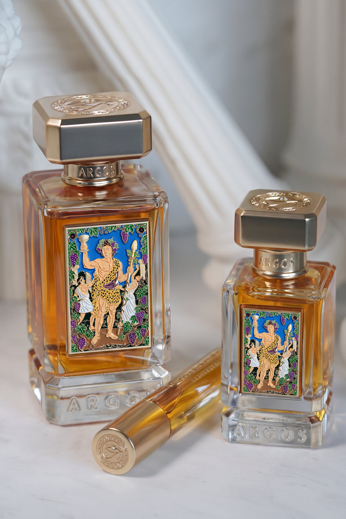 Two argos la dolce vita di bacchus perfume bottles with ornate gold and multicolor labels, one large and one small, with a matching gold atomizer tube on a white marble surface.
