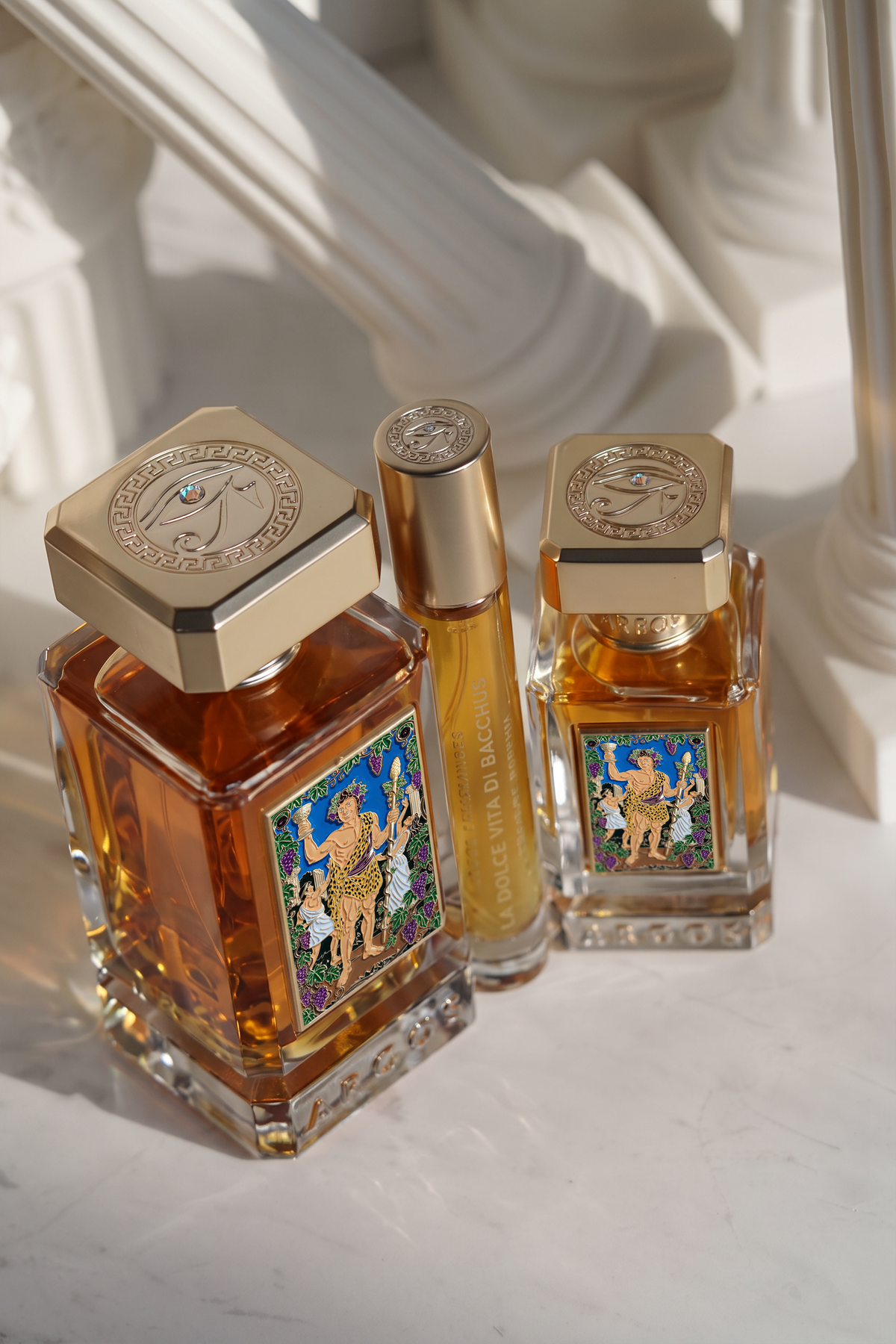 Three perfume bottles from argos la dolce vita di bacchus displayed on a light marble surface with classical white columns in the background. The set includes a large rectangular bottle with a gold cap, a matching small rollerball bottle, and a smaller rectangular bottle, all filled with amber-colored fragrance and featuring colorful illustrated labels.