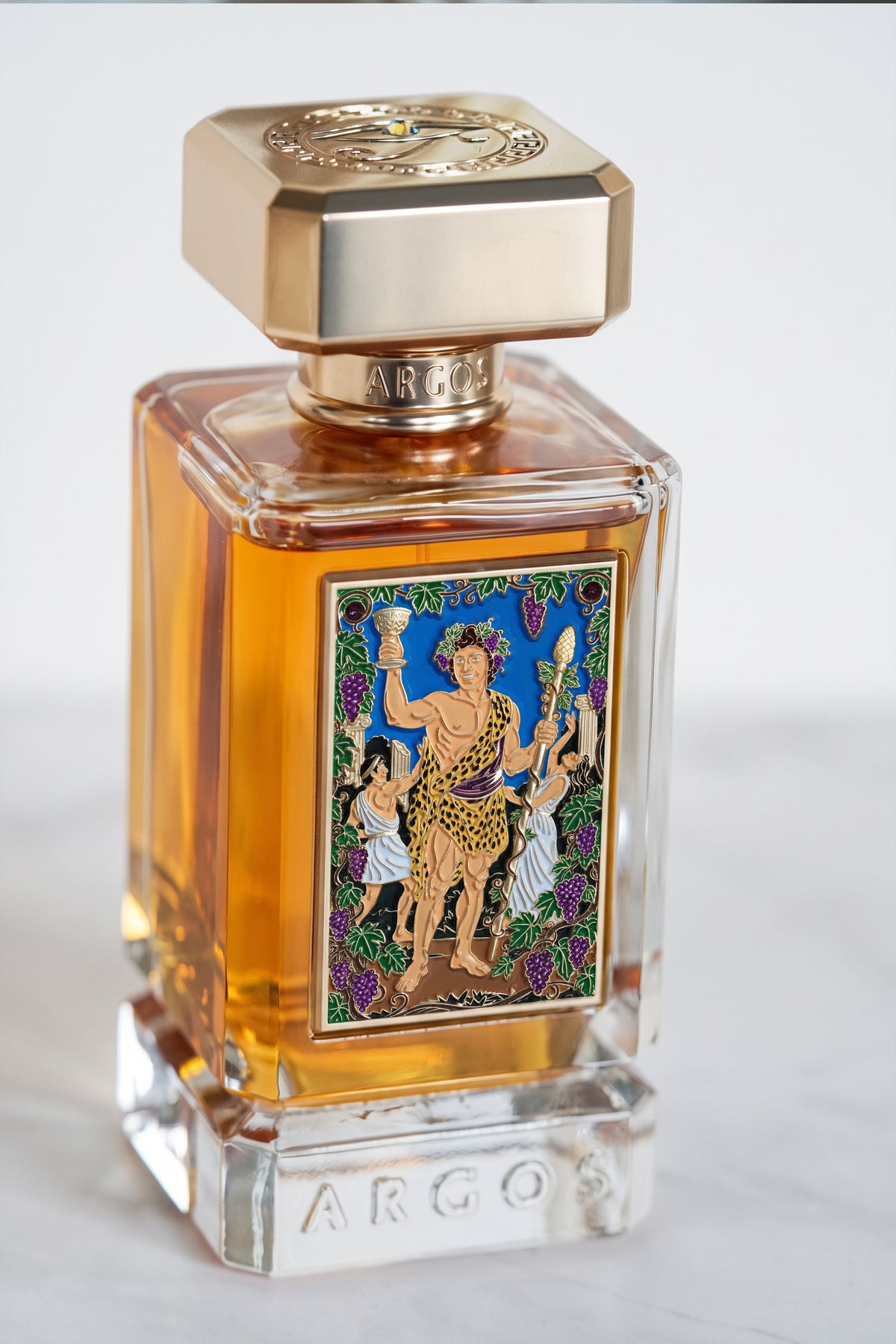 argos la dolce vita di bacchus 100ml perfume bottle with amber liquid, a gold-toned cap, and a colorful illustrated front label, photographed on a light neutral background.
