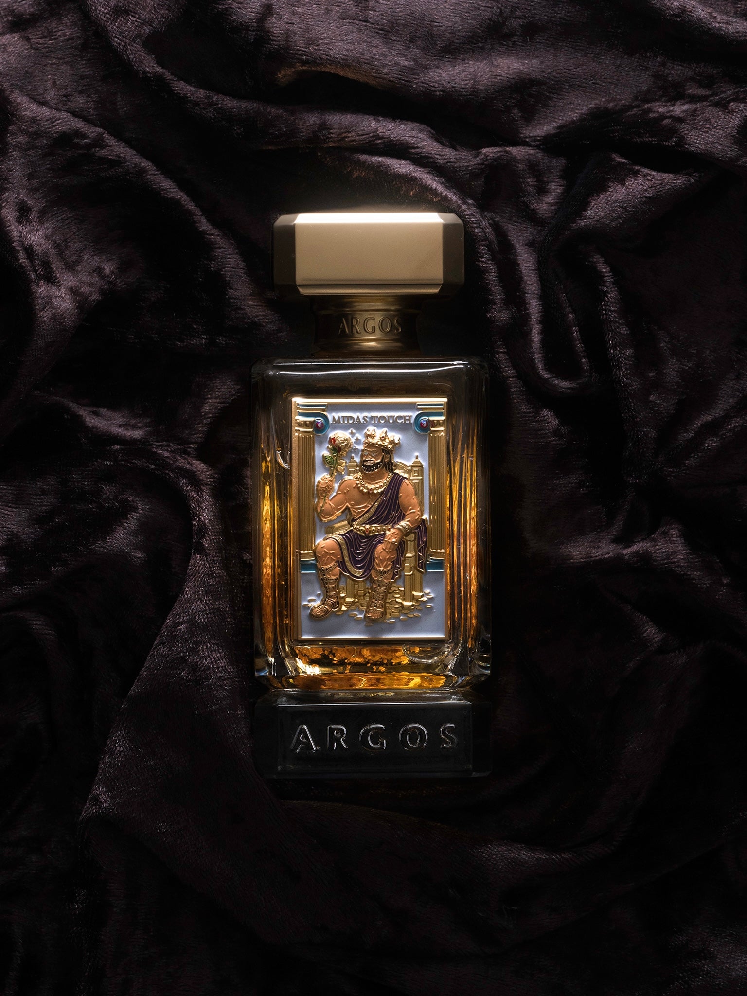 Gold and silver Argos Midas Touch 24K Edition 100ml perfume bottle on a dark fabric background